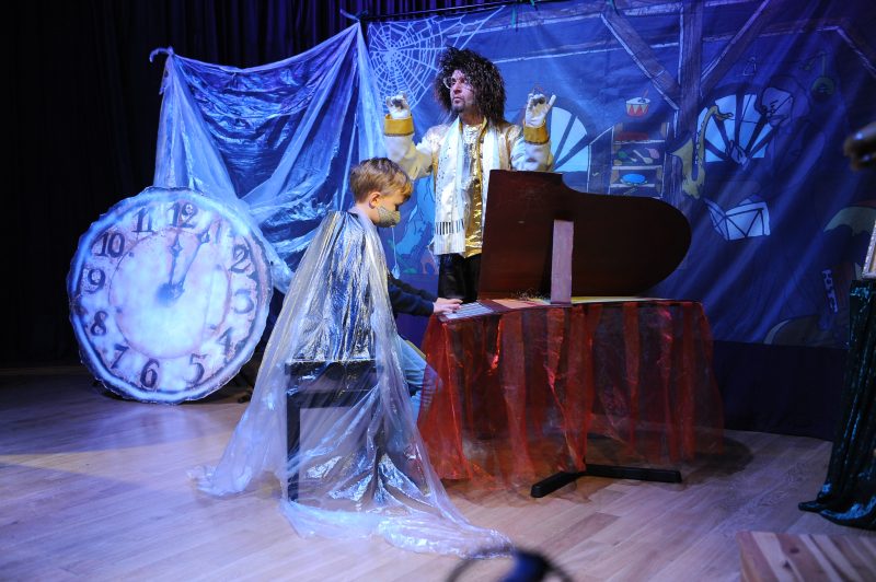 Chłopiec w pelerynie gra na fortepianie, aktor dyryguje, tarcza starego zegara