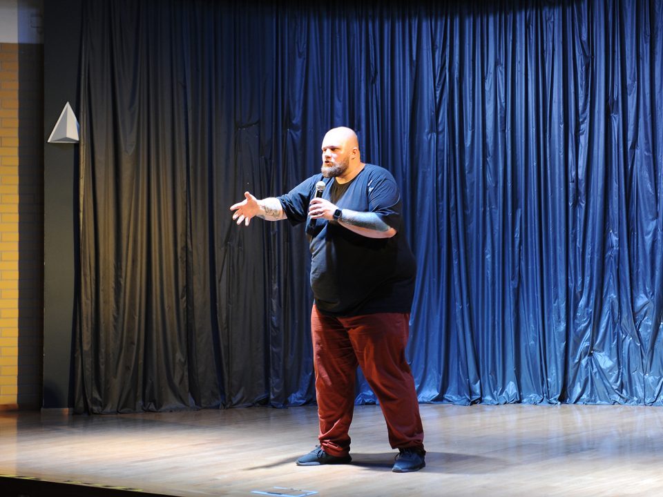 Mężczyzna z brodą na scenie gestykuluje ręką mówiąc do mikrofonu